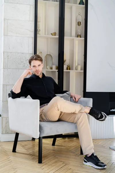 Вид в полный рост стильного мужчины в черной рубашке и бежевых брюках, сидящего в кресле — стоковое фото