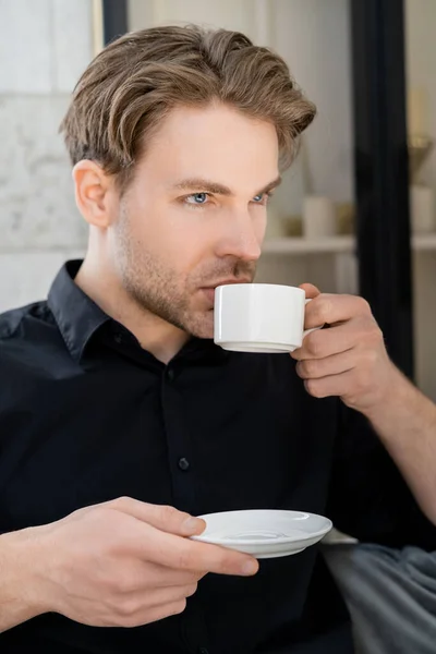 Homme en chemise noire tenant soucoupe et boire du café de tasse blanche — Photo de stock