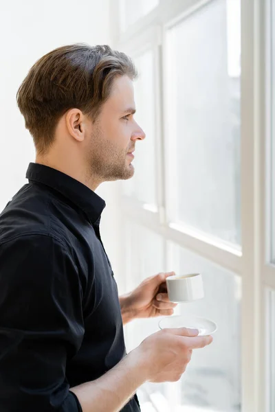 Vista lateral do homem em camisa preta de pé com xícara de café perto da janela — Fotografia de Stock