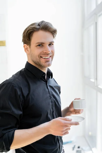 Hombre feliz en camisa negra sosteniendo taza de café y sonriendo a la cámara - foto de stock
