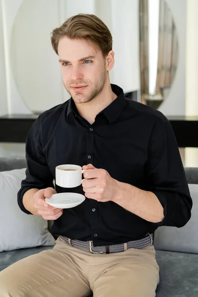 Mann im schwarzen Hemd sitzt mit Kaffeetasse und Untertasse auf Couch im Wohnzimmer — Stockfoto