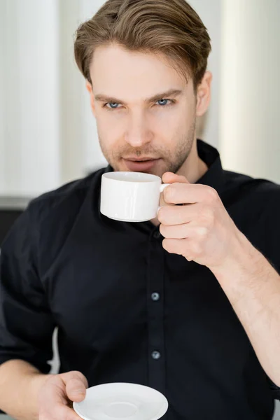 Молодий чоловік у чорній сорочці п'є ранкову каву з білої чашки — стокове фото