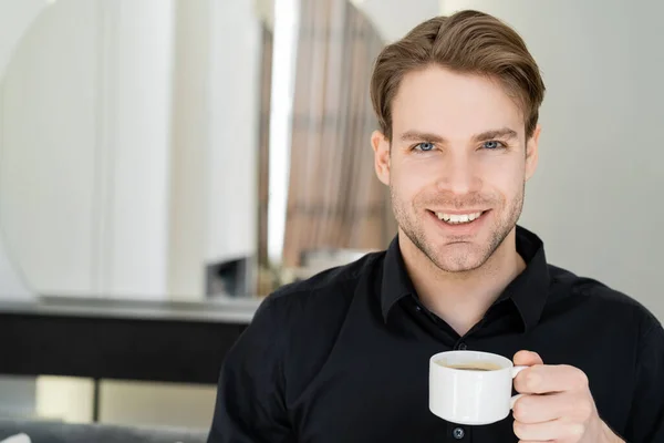 Mann im schwarzen Hemd hält weiße Kaffeetasse in der Hand und lächelt in die Kamera — Stockfoto