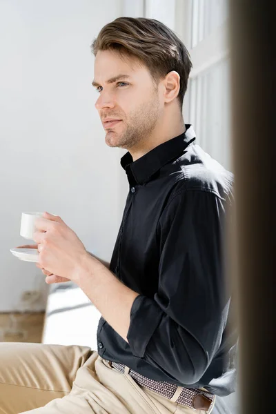 Hombre de camisa negra sosteniendo la taza de café mientras está sentado y mirando hacia otro lado en casa - foto de stock