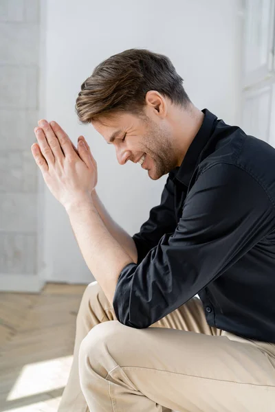 Vista lateral del hombre con las manos orantes sonriendo con los ojos cerrados en casa - foto de stock