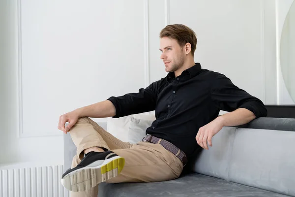 Man in black shirt and beige trousers sitting on sofa in living room — Stock Photo