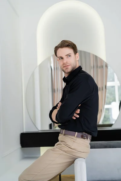 Uomo in camicia nera seduto con le braccia incrociate vicino allo specchio sfocato e distogliendo lo sguardo — Foto stock