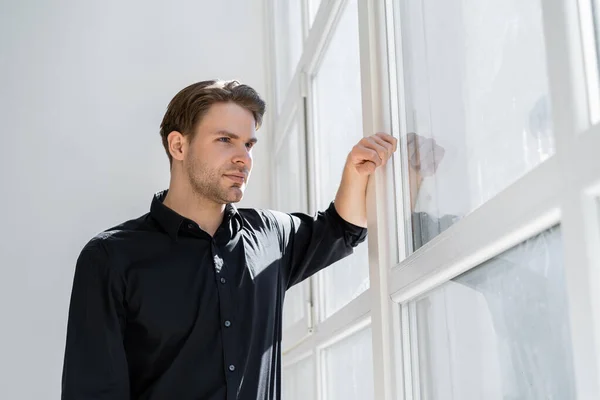 Nachdenklicher junger Mann in schwarzem Hemd steht am Fenster und schaut weg — Stockfoto