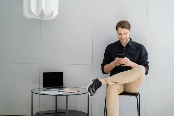 Homme heureux tapant sur smartphone tout en étant assis avec les jambes croisées près de l'ordinateur portable avec écran vide — Photo de stock