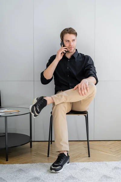 Vista completa del hombre elegante sentado cerca de la pared gris y hablando en el teléfono inteligente - foto de stock