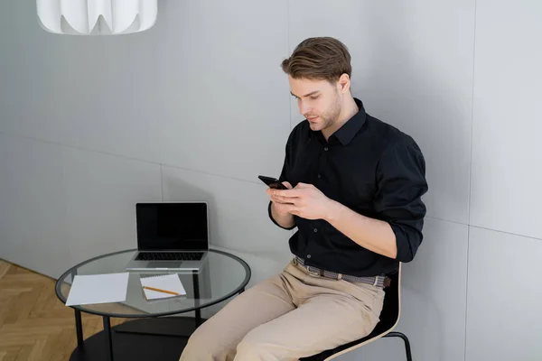 Человек сидит рядом с ноутбуком с пустым экраном на кофейном столике и обмена сообщениями на смартфоне — стоковое фото