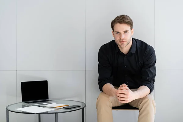 Junger Mann blickt in die Kamera, während er neben Laptop mit leerem Bildschirm an grauer Wand sitzt — Stockfoto