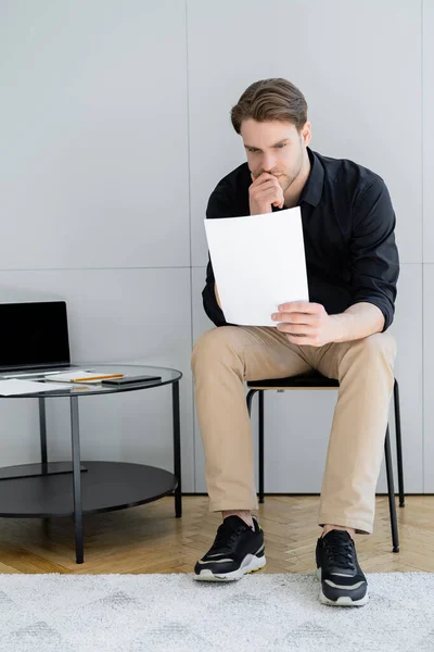 Longitud completa del hombre reflexivo mirando el documento mientras está sentado cerca de la computadora portátil con pantalla en blanco - foto de stock