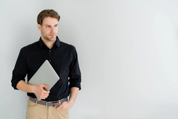 Uomo in camicia nera che tiene il computer portatile in piedi con mano in tasca isolato su grigio — Foto stock