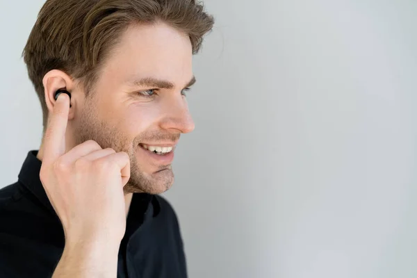 Uomo sorridente regolazione auricolare wireless durante l'ascolto di musica isolata su grigio — Foto stock