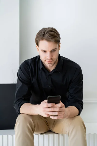 Стильний чоловік в чорній сорочці, сидячи вдома і обмін повідомленнями на смартфоні — стокове фото