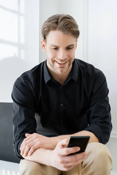 Uomo allegro in camicia nera utilizzando il telefono cellulare a casa — Foto stock