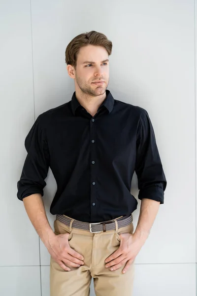 Hombre con estilo en camisa negra y pantalones beige de pie con pulgares en bolsillos cerca de la pared gris - foto de stock