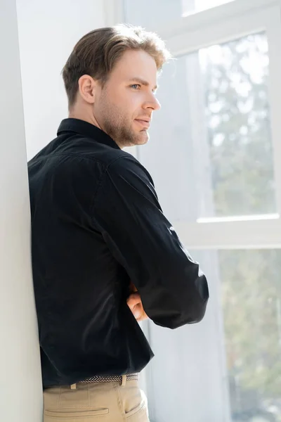 Giovane uomo in camicia nera in piedi vicino alla finestra e guardando altrove — Foto stock