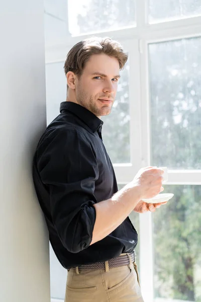 Young man standing near window with coffee cup and looking at camera — Stock Photo