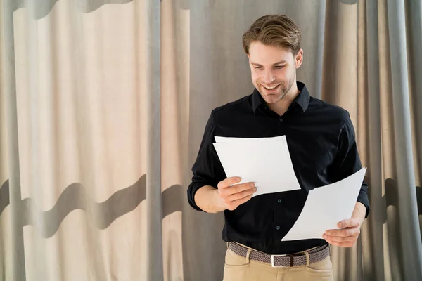 Hombre joven y positivo en camisa negra mirando papeles cerca de cortina beige - foto de stock