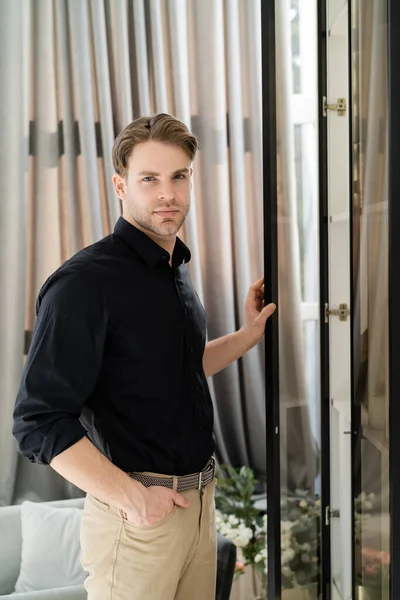 Homme en chemise noire debout avec la main dans la poche près de la fenêtre et rideau beige à la maison — Photo de stock