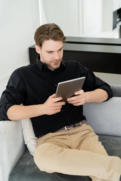 Junger Mann in schwarzem Hemd sitzt zu Hause auf Couch und nutzt digitales Tablet — Stockfoto