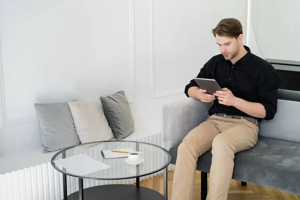 Молодой человек с помощью цифрового планшета рядом с журнальным столиком в гостиной — стоковое фото