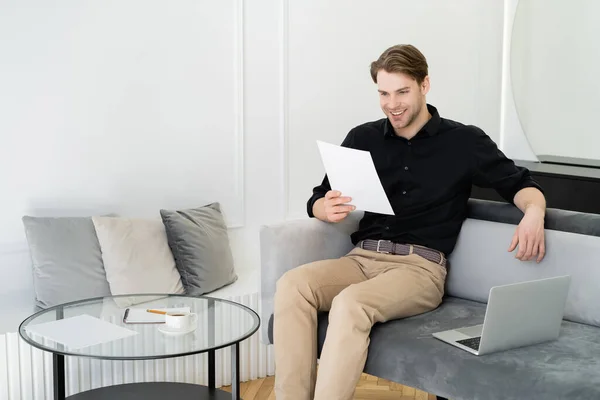 Alegre hombre leyendo documento mientras está sentado en el sofá cerca de la computadora portátil y mesa de café - foto de stock