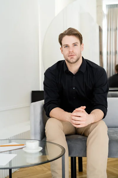 Mann im schwarzen Hemd blickt in die Kamera, während er auf dem Sofa am Couchtisch sitzt — Stockfoto