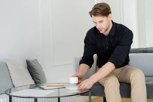 Junger Mann im schwarzen Hemd mit einer Tasse Kaffee auf der Couch sitzend — Stockfoto