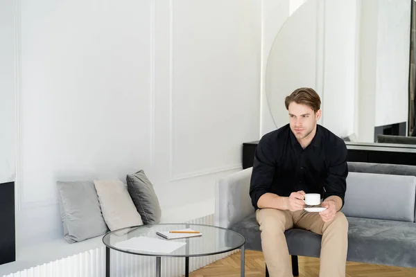 Young man in black shirt sitting with cup near coffee table — Stock Photo