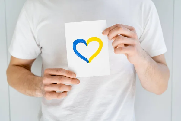 Partial view of man in white t-shirt holding card with blue and yellow heart on grey background — Stock Photo