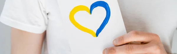 Tarjeta blanca con corazón azul y amarillo en la mano del hombre recortado aislado en gris, bandera - foto de stock