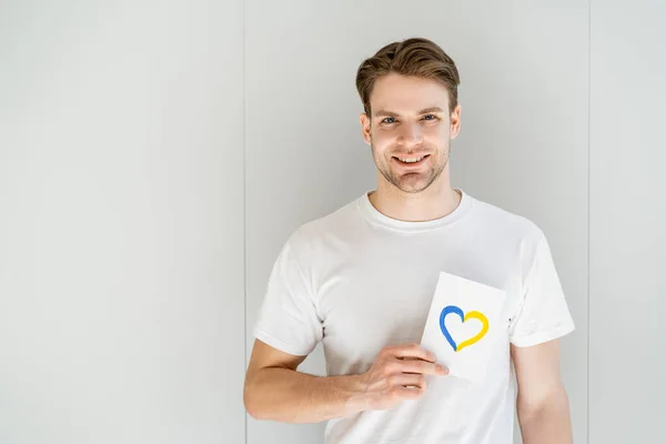 Hombre feliz sosteniendo la tarjeta blanca con corazón azul y amarillo sobre fondo gris — Stock Photo