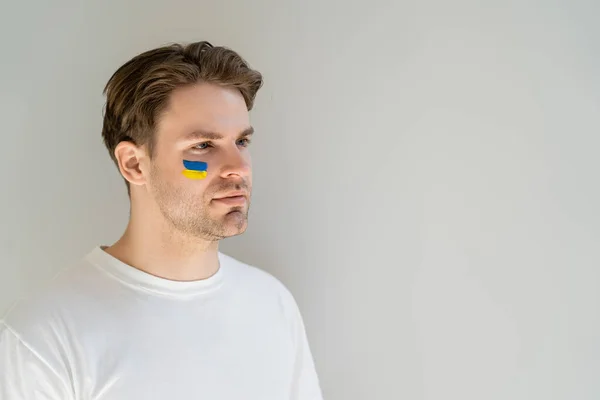 Joven patriota con bandera ucraniana pintada en la cara mirando hacia otro lado aislado en gris - foto de stock