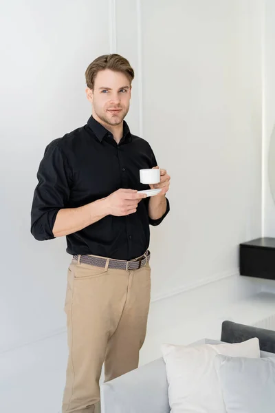 Hombre joven en camisa negra y pantalones beige de pie con taza de café y platillo en la sala de estar - foto de stock