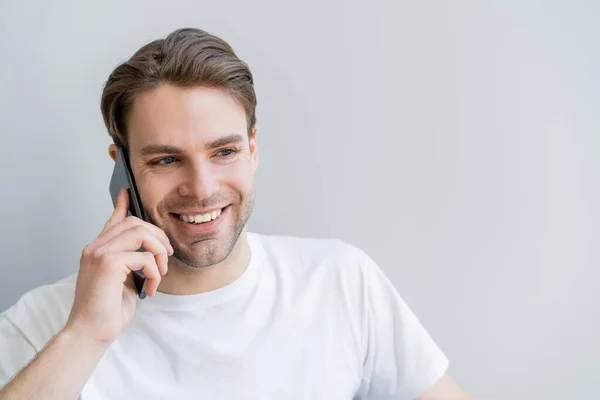 Щасливий молодий чоловік у білій футболці говорить на смартфоні ізольовано на сірому — стокове фото