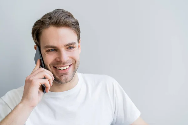 Heureux jeune homme en t-shirt blanc parlant sur smartphone isolé sur gris — Photo de stock