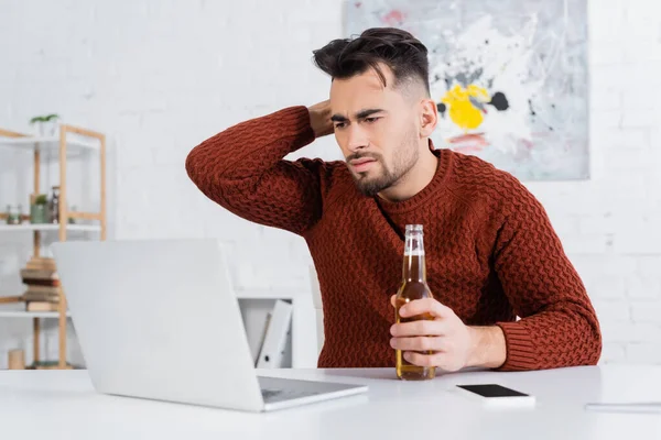 Displeased bookmaker with bottle of beer looking at blurred laptop - foto de stock