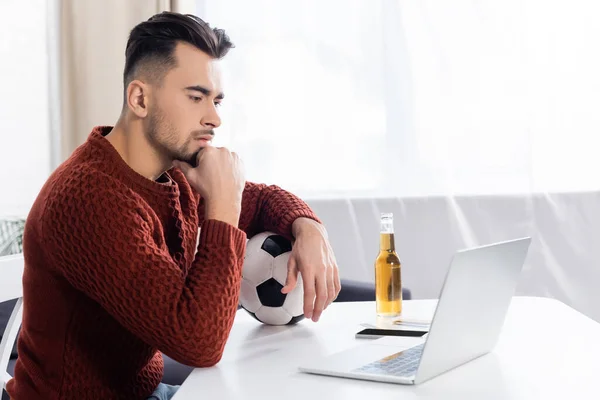 Casa de apuestas enfocada con pelota de fútbol mirando el portátil cerca de la botella de cerveza - foto de stock