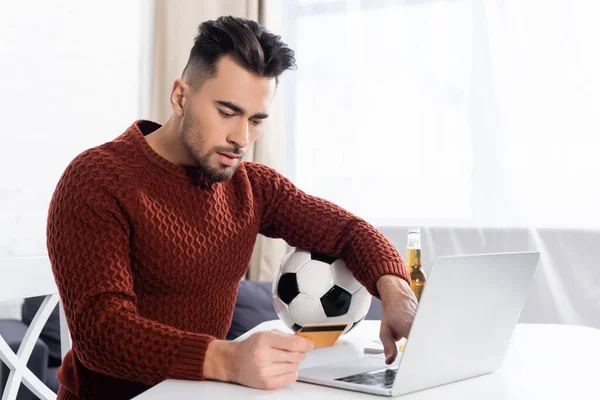 Young bookmaker holding credit card near laptop and soccer ball — Photo de stock