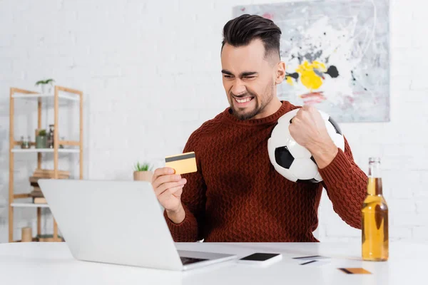 Взволнованный букмекер с кредитной картой и футбольный мяч возле ноутбука и бутылки пива — стоковое фото