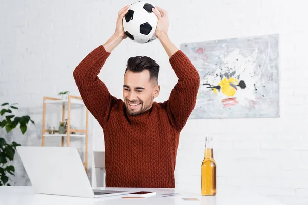 Joyful bookmaker holding soccer ball above head near laptop and beer — Foto stock
