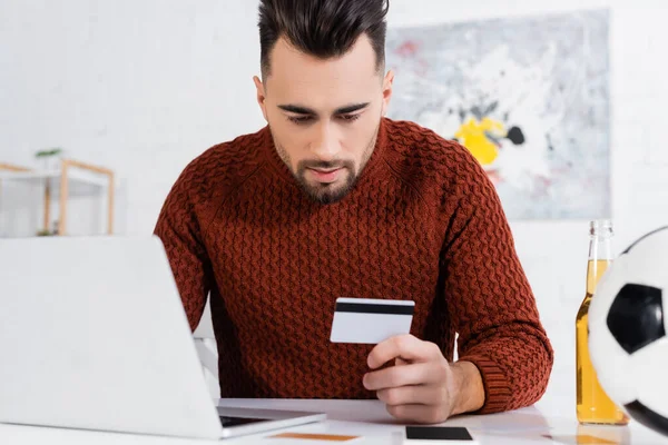 Serious bookmaker holding credit card near laptop and blurred soccer ball — Photo de stock