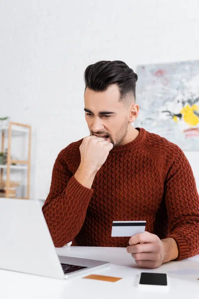 Nervous bookmaker biting fist while holding credit card near laptop — Stock Photo