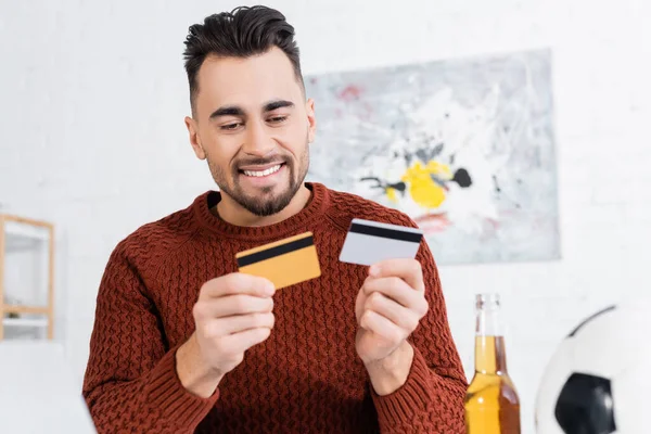 Sorrindo bookmaker segurando cartões de crédito perto bola de futebol borrada e cerveja — Fotografia de Stock