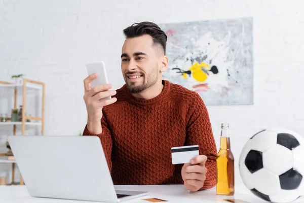 Joueur souriant avec carte de crédit regardant smartphone près d'un ordinateur portable, bière et ballon de football — Photo de stock