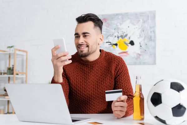 Smiling gambler with credit card looking at smartphone near laptop, beer and soccer ball — Stock Photo
