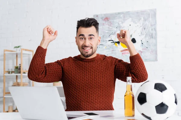 Aufgeregter Buchmacher zeigt Erfolgsgeste in der Nähe von Laptop, Bier und Fußball — Stockfoto